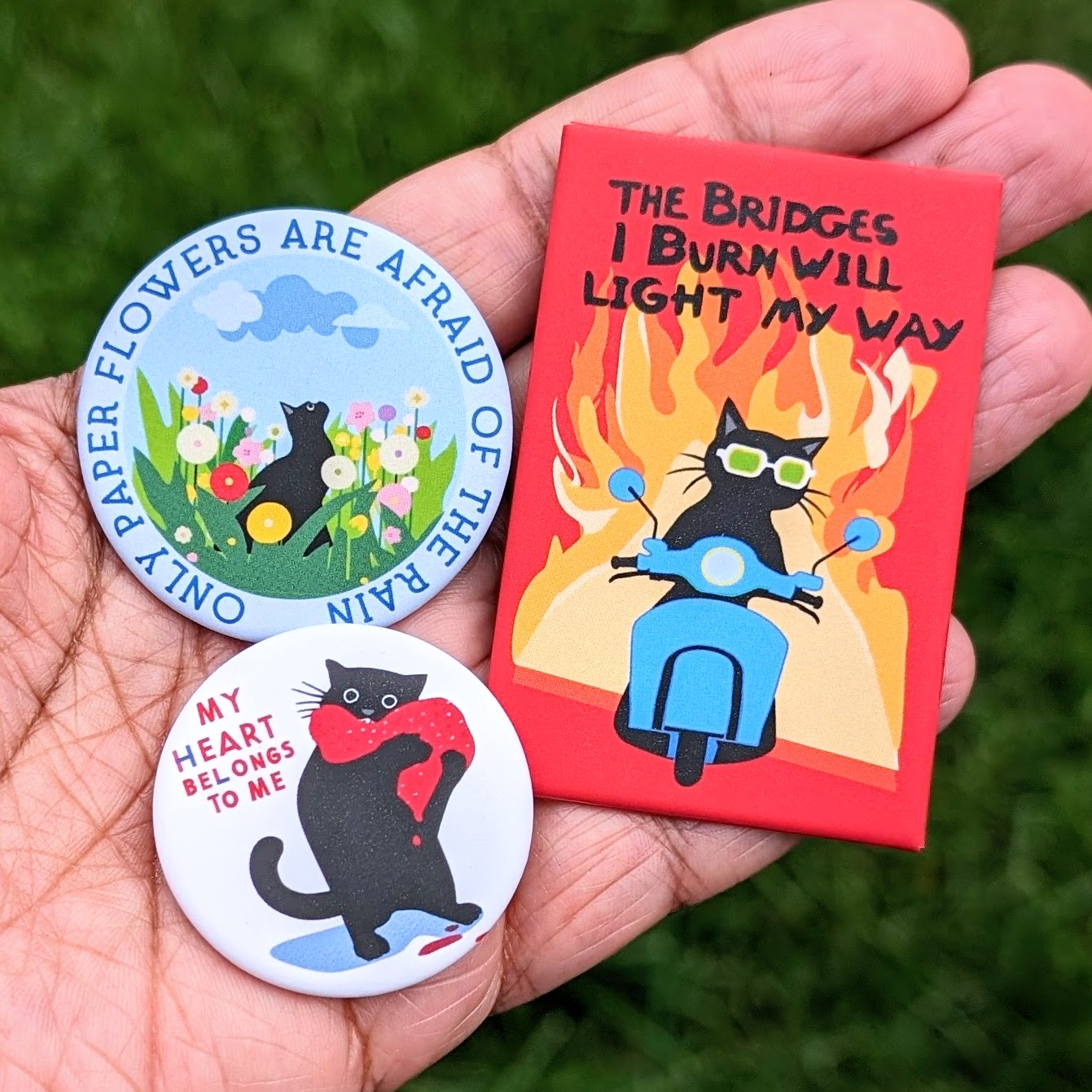 3 pin back buttons with designs that feature black cats. One buttons says: The bridges I burn will light my way; 2nd button says: Only paper flowers are afraid of the rain; 3rd button says: My heart belongs to me